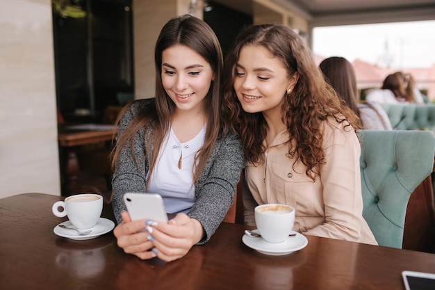 Duas amigas bebem café no café e olham no telefone inteligente Mulheres sorridentes felizes sentadas no terraço Pessoas olham histórias