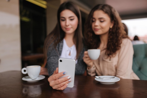 Duas amigas bebem café no café e olham no telefone inteligente Mulheres sorridentes felizes sentadas no terraço Pessoas olham histórias