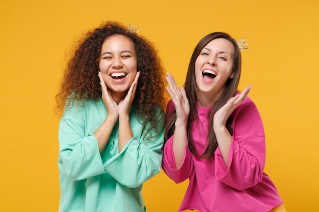 Duas amigas bonitas garotas europeias e afro-americanas em roupas verdes rosa, coroas posando isoladas no fundo da parede amarela. Conceito de estilo de vida de pessoas. Mock up espaço de cópia. Coloque as mãos nas bochechas.