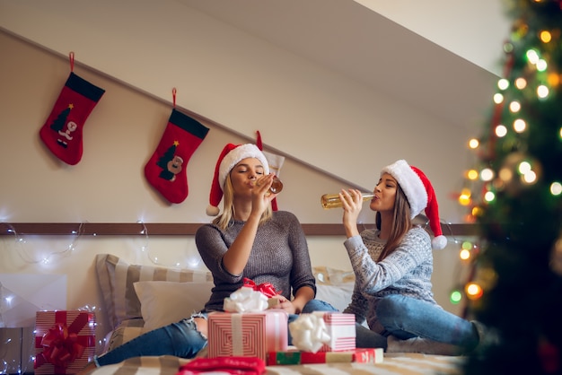Duas amigas brincalhonas com chapéu de Papai Noel sentadas na cama para as férias de Natal e comemorando bebendo cerveja