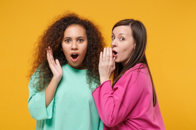 Duas amigas chocadas afro-americanas europeias em roupas verdes rosa isoladas em fundo amarelo. Conceito de estilo de vida de pessoas. Mock up espaço de cópia. Sussurrando segredo por trás da mão, espalhando as mãos.