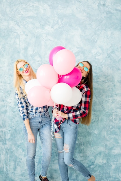 Duas amigas com camisas xadrez se divertindo com balões de ar no fundo da parede azul