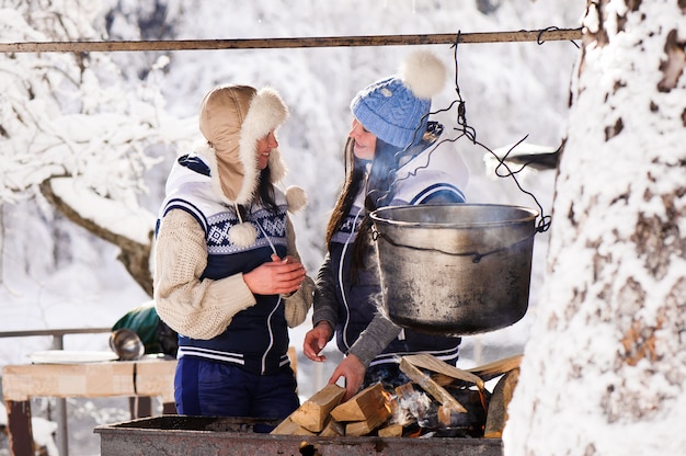 Duas amigas cozinhando na natureza inveja em um incêndio na caldeira. Meninas aquecer no fogo no inverno.