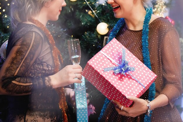 Duas amigas dão caixas de presente uma à outra perto de uma árvore de Natal.