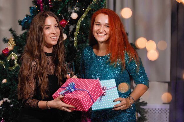 Duas amigas dão caixas de presente uma à outra perto de uma árvore de Natal.