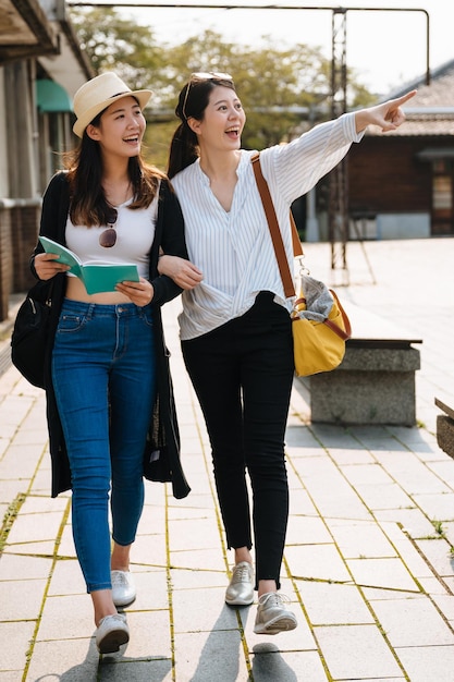 duas amigas de viagens chinesas asiáticas andando pela área da cidade velha nas férias de verão com o livro guia se divertindo. amigos de mulheres jovens rindo de corpo inteiro mostrando de lado enquanto relaxam o dia ensolarado ao ar livre