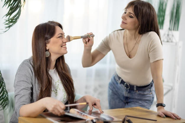 Duas amigas em roupas elegantes fazem maquiagem uma à outra perto da mesa com uma revista