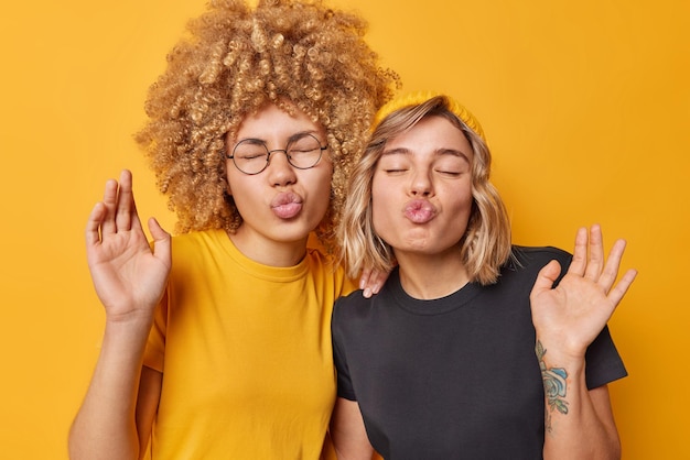 Duas amigas engraçadas se divertem lábios de beicinho mantém os olhos fechados vestidos com camisetas casuais tolas por aí fazer careta isolada sobre fundo amarelo brilhante amizade e conceito de tempo livre