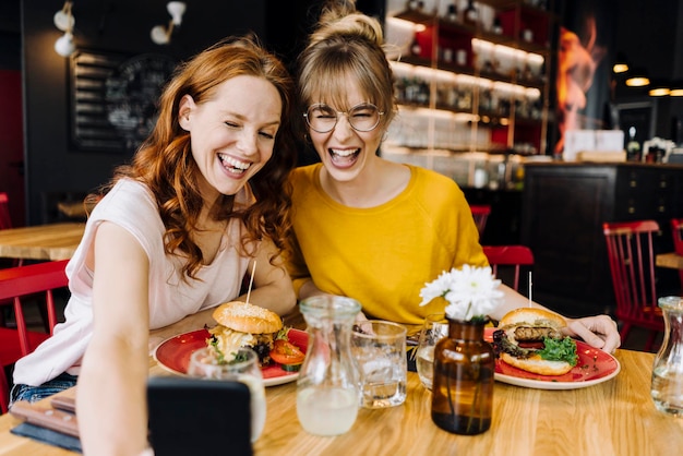Duas amigas felizes comendo hambúrguer e tirando uma selfie em um restaurante.