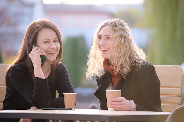 Duas amigas felizes se divertindo juntas no café de rua rindo e falando no celular