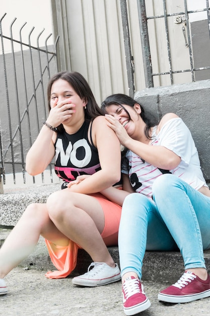 duas amigas latinas sentadas na rua, conversando e rindo alto, cobrindo a boca