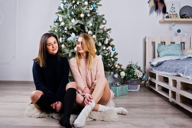 Duas amigas lindas usam camisola quente e polainas de polainas contra árvore de ano novo com decoração de natal Retrato de férias de estúdio de menina loira e morena