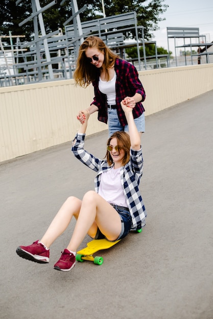 Duas amigas sorridentes se divertindo montando longboard amarelo na rua. Conceito de amizade.