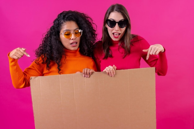 Duas amigas sorrindo e apontando para a placa de papelão em um fundo rosa