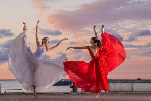 Duas bailarinas em uma saia voadora branca e vermelha e collant dançando em dueto na margem do oceano ou mar contra o pano de fundo do céu pôr do sol