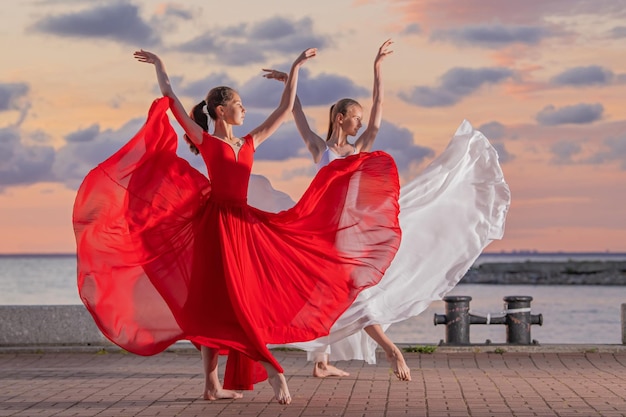 Duas bailarinas em uma saia voadora branca e vermelha e collant dançando em dueto na margem do oceano ou mar contra o pano de fundo do céu pôr do sol