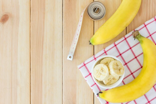 Duas bananas e fatias de banana em uma tigela branca com fita métrica na mesa de madeira