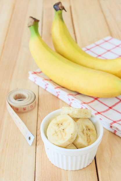 Duas bananas e fatias de banana em uma tigela branca com fita métrica na mesa de madeira