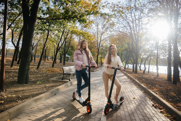 Duas belas garotas andam de scooters elétricas no parque em um dia quente de outono.