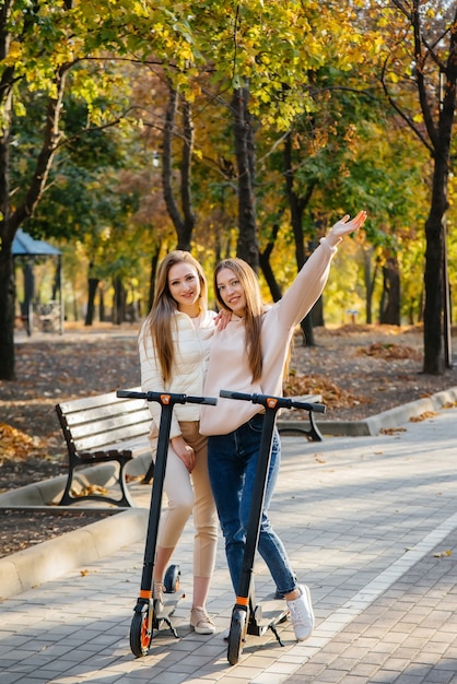 Duas belas garotas andam de scooters elétricas no parque em um dia quente de outono.