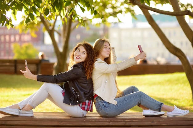 Duas belas moças tiram selfies em um parque ensolarado ao mesmo tempo. Amigas.