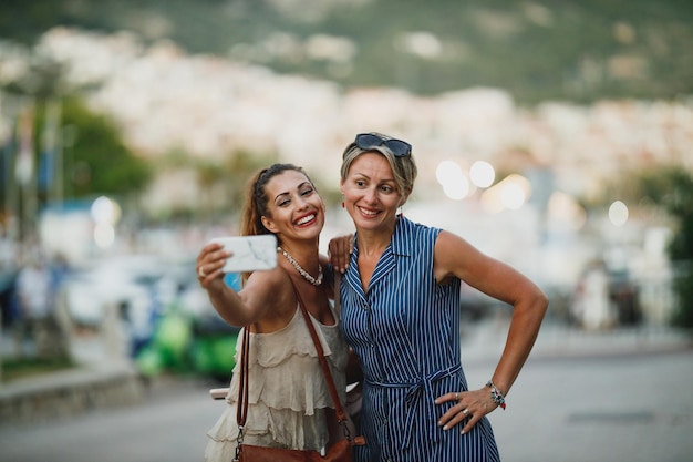 Duas belas mulheres estão se divertindo e tirando uma selfie enquanto caminham pela rua de uma cidade mediterrânea.