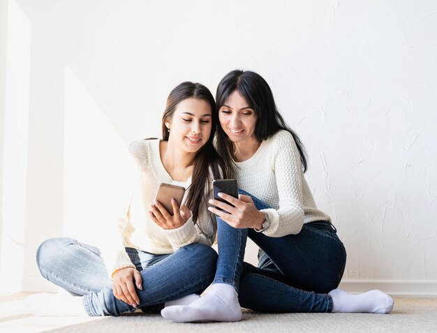 Duas belas mulheres rindo de jeans e suéteres brancos enviando mensagens de texto para amigos usando telefones celulares