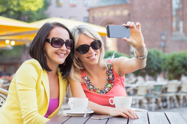 Duas belas mulheres tirando fotos de selfie com o celular enquanto estão sentadas no café ao ar livre