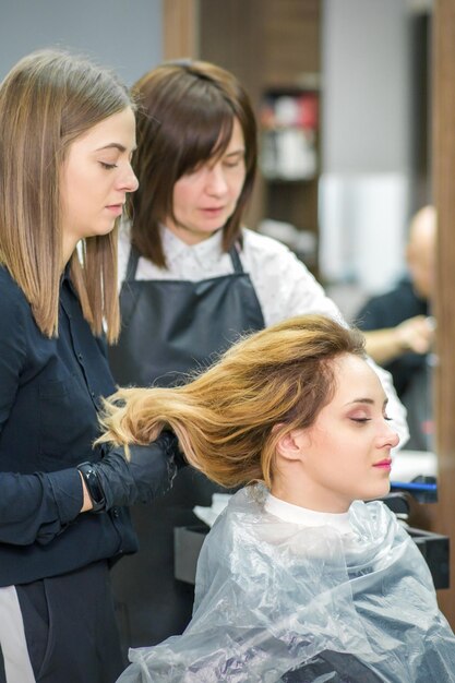 Duas cabeleireiras femininas preparam o cabelo comprido de uma jovem mulher fazendo penteado cachos em um salão de beleza.