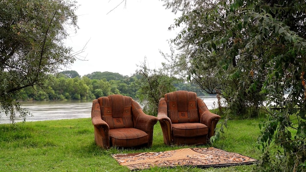 Duas cadeiras estão nas margens do rio, há árvores ao redor e um tapete na frente