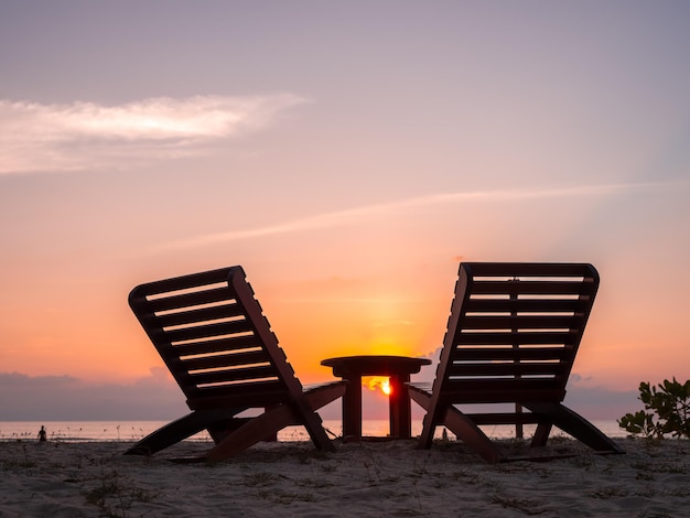 Duas camas de sol vazias de silhueta de madeira, cadeira de casal e mesa lateral na praia de areia, fundo de céu tranquilo ao pôr-do-sol à noite com cópia de espaço, conceito de férias ou férias de verão