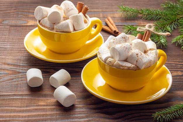 Duas canecas de cerâmica amarela com café com leite com marshmallows e canela ao lado de ramos de abeto