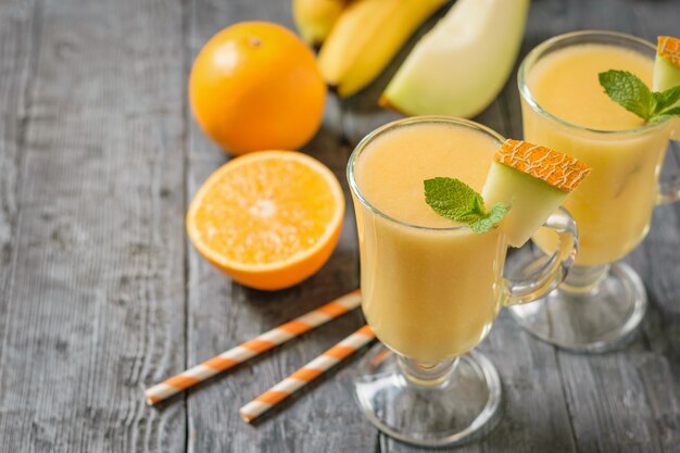 Duas canecas de vidro com suco de melão, laranja e melão em uma mesa de madeira preta.