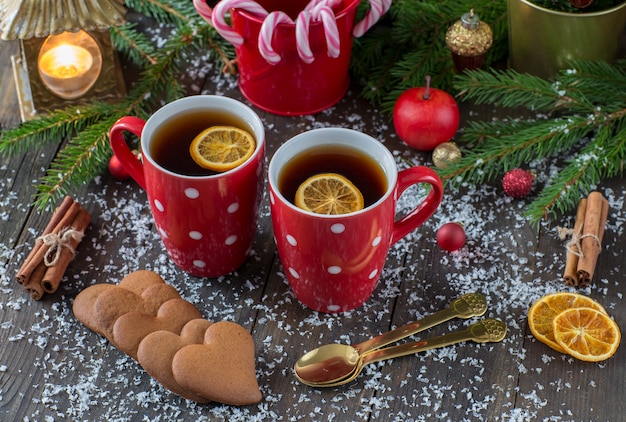 Duas canecas vermelhas com chá, cana caramelo, biscoitos em forma de coração, uma maçã, uma lanterna