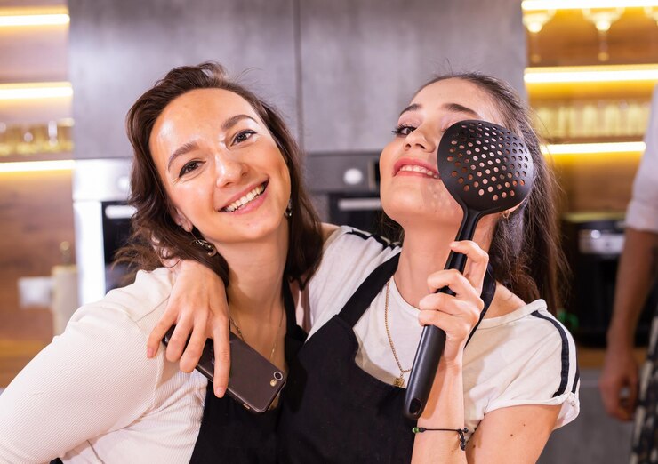Foto duas chefs engraçadas de uniforme segurando utensílios de cozinha enquanto sorriem e se divertem com a câmera na cozinha