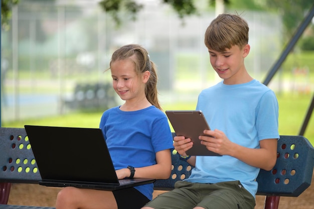 Duas crianças adolescente menino com computador portátil e jovem com tablet digital olhando para telas ao ar livre sentado no banco no parque de verão Educação remota durante o conceito de quarentena