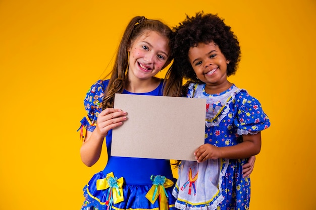 Duas crianças amigas com roupa de festa junina segurando um cartão em branco com espaço de cópia