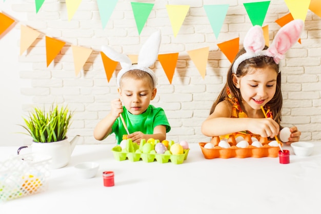 Duas crianças com orelhas de coelho da Páscoa pintando ovos de Páscoa coloridos em casa. O menino e a menina estão se preparando para a Páscoa