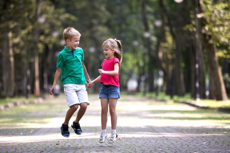 Foto duas crianças de sorriso engraçadas novas bonitos, menina e menino, irmão e irmã, saltando e se divertindo no bokeh ensolarado borrado das árvores do verde da aleia do parque ensolarado. conceito de infância descuidada feliz.