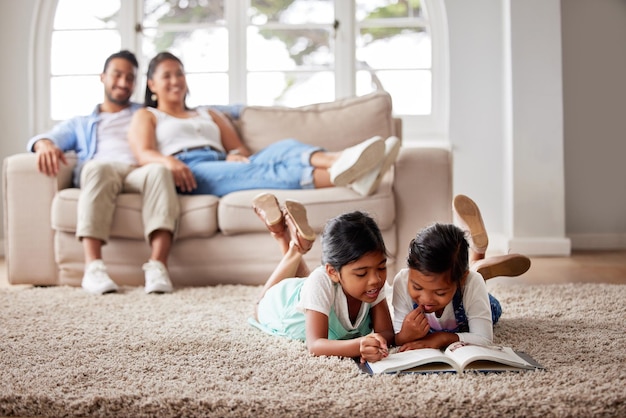 Duas crianças lendo um livro no chão da sala enquanto seus pais relaxam em um sofá ao fundo em casa.