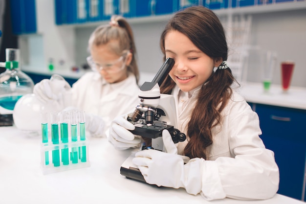 Duas crianças no jaleco aprendendo química no laboratório da escola. Jovens cientistas em óculos de proteção fazendo experimento em laboratório ou gabinete de químico. Olhando através do microscópio