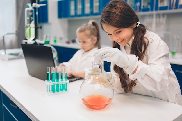 Duas crianças no jaleco aprendendo química no laboratório da escola. jovens cientistas em óculos de proteção fazendo experimento em laboratório ou gabinete de químico. trabalhando em um pc