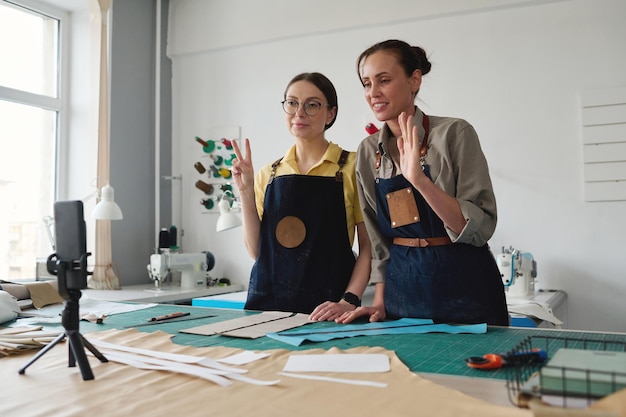 Duas curtidoras femininas criativas em roupas de trabalho cumprimentando o público on-line