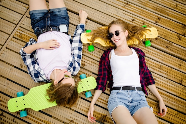 Duas elegantes jovens urbanas com longboards deitam no chão de madeira na rua. Os amigos se divertem e passam tempo juntos no verão durante as férias.