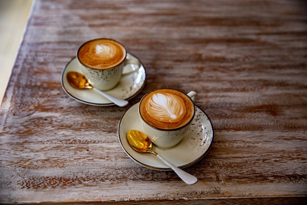 Duas elegantes xícaras brancas de cappuccino com latte art no pires e colheres de ouro na mesa de madeira clara. Bebida de café da manhã