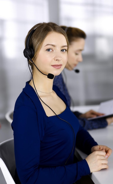 Duas empresárias conversam com os clientes por meio de fones de ouvido, enquanto estão sentadas à mesa de um escritório moderno. Grupo de pessoas diversas em um call center. Telemarketing e atendimento ao cliente.