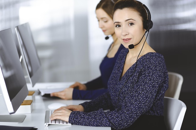 Duas empresárias conversam com os clientes por meio de fones de ouvido, enquanto estão sentadas à mesa de um escritório moderno. Grupo de pessoas diversas em um call center. Telemarketing e atendimento ao cliente.