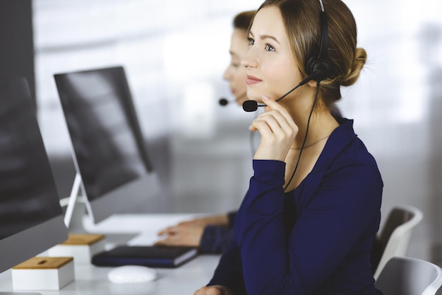 Duas empresárias conversam com os clientes por meio de fones de ouvido, enquanto estão sentadas à mesa de um escritório moderno. Grupo de pessoas diversas em um call center. Telemarketing e atendimento ao cliente.