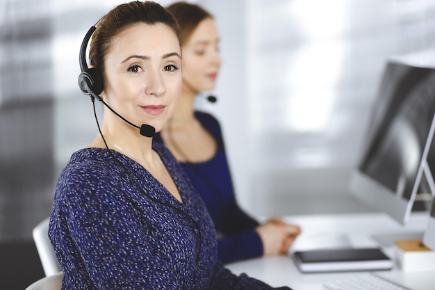 Duas empresárias conversam com os clientes por meio de fones de ouvido, enquanto estão sentadas à mesa de um escritório moderno. Grupo de pessoas diversas em um call center. Telemarketing e atendimento ao cliente.