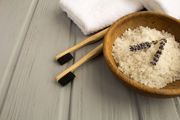 Duas escovas de dente de bambu, toalha branca e sal de banho marinho na tigela marrom sobre o fundo cinza de madeira